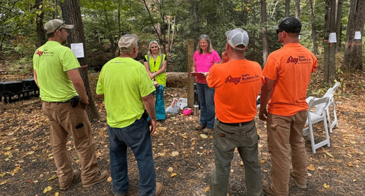 Group of people in neon shirts standing in a circle in a forest, with a woman speaking and papers posted on trees in the background.