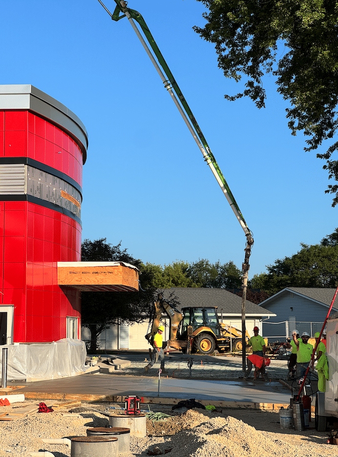Screenshot 2026-07-23 at 32135 PM | Holtz Builders | General Contractor Construction site with a red modern building and a long concrete pump arm pouring cement into a foundation, workers in neon vests nearby.