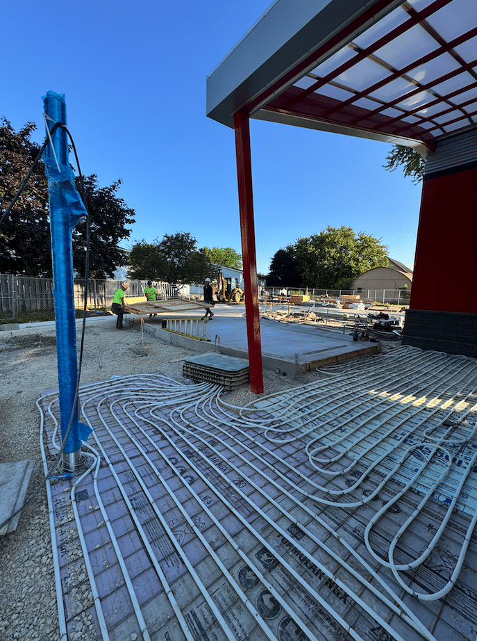 Tommys Car Wash BD | Holtz Builders | General Contractor Construction site showing radiant floor heating tubes laid in a grid on a concrete slab; blue-wrapped pipe in foreground and a red steel column, with workers in the distance.